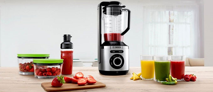 یک مخلوط کن bosch نقرهای و مشکی در کنار سه لیوان آبمیوه، دو ظرف میوه پر از توت فرنگی و یک بطری آبمیوه