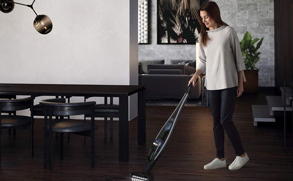 زنی با لباس روزمره در یک اتاق پذیرایی با کفپوش چوبی در حال جارو زدن با جارو شارژی برند AEG است.